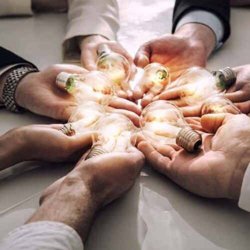 A bunch of people holding laying their hands out in a circle, each with a lit light bulb in it.