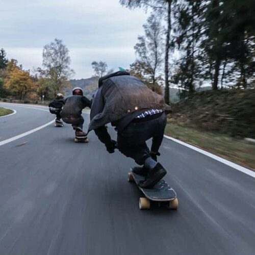 Three people skating on skateboards downhill with motion blue showing immense speed, surrounded by trees and hills.